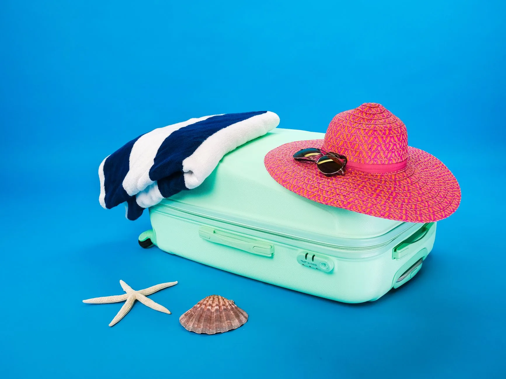 Türkiser Koffer am Strand mit Sonnenhut, Sonnenbrille und Muscheln